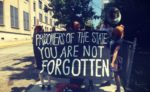 People outside a prison with a musical instruments and a banner reading "Prisoners of the state: You Are Not Forgotten"