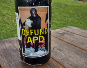 "Defund APD" sticker on a water bottle, depicting an asheville police officer stabbing and crushing water bottles after raiding a medic table during George Floyd protests in 2020