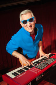 Image description: photograph of Keith Munslow wearing sunglasses & smiling with his hands on a keyboard. He is a white man with white hair & a goatee.
